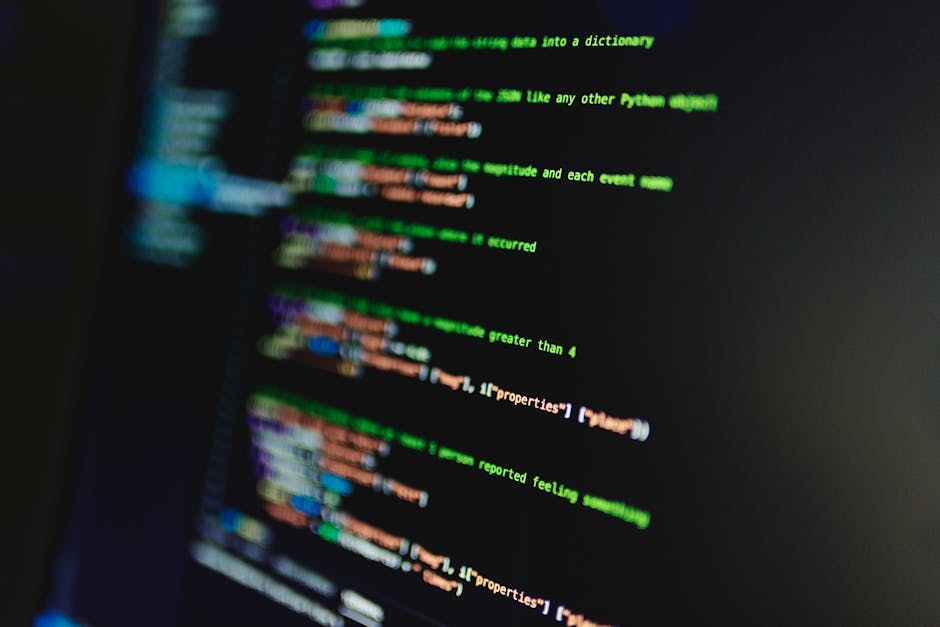 Close-up view of a computer screen displaying lines of colorful programming code in green, orange, blue, and purple on a dark background, with a blurred effect focusing on the text. The screen appears